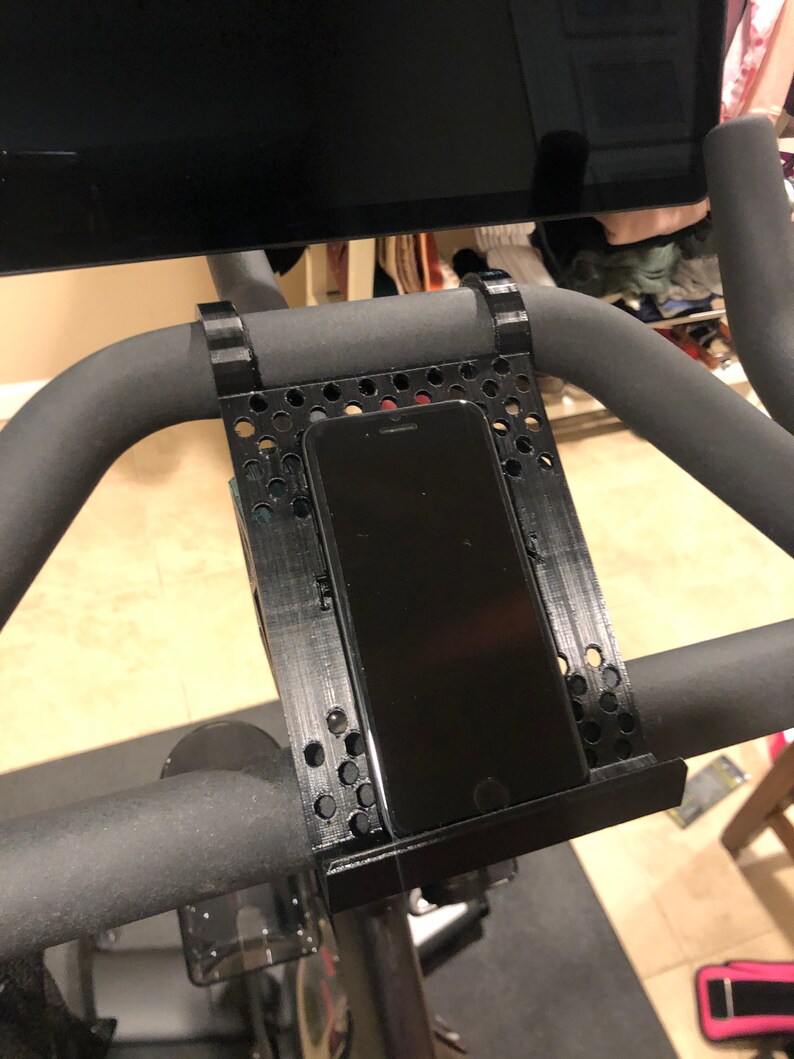 May include: Black phone holder mounted on the handlebars of a stationary exercise bike. The holder is made of black plastic and has a grid pattern. The phone is in a vertical position.