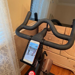 May include: A black stationary exercise bike with a tablet mounted on the handlebars. A smartphone is mounted on the handlebars in a phone holder. The phone screen shows a fitness app with a timer displaying 1:30.
