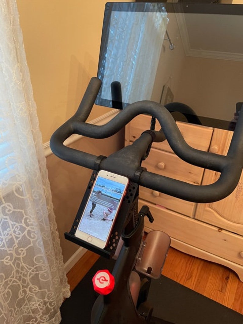 May include: A black stationary exercise bike with a tablet mounted on the handlebars. A smartphone is mounted on the handlebars in a phone holder. The phone screen shows a fitness app with a timer displaying 1:30.