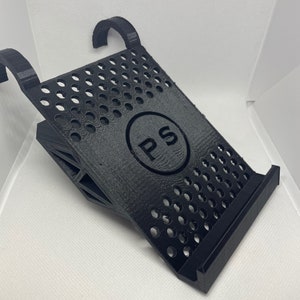 May include: Black 3D printed stand with a circular cutout featuring the letters "PS" in a circle. The stand has a grid pattern and two hooks on the top.