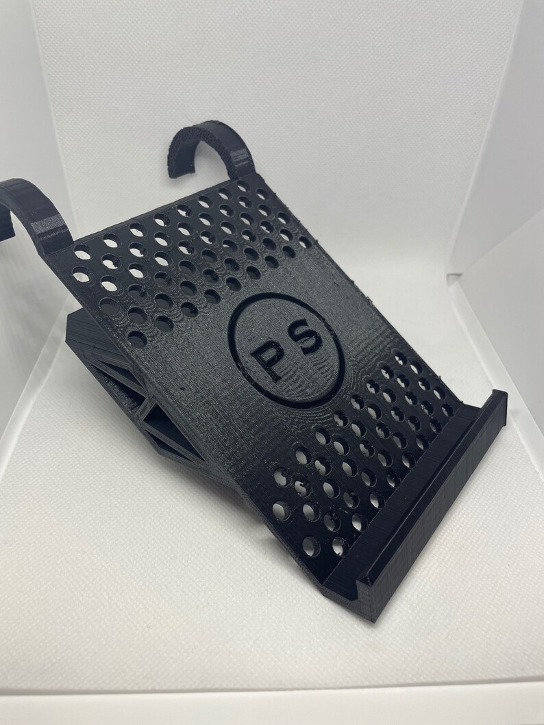 May include: Black 3D printed stand with a circular cutout featuring the letters "PS" in a circle. The stand has a grid pattern and two hooks on the top.