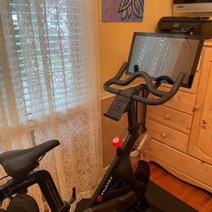 May include: A black stationary exercise bike with a touchscreen display and a red "Peloton" logo. The bike is in a home setting with a window and a dresser in the background.