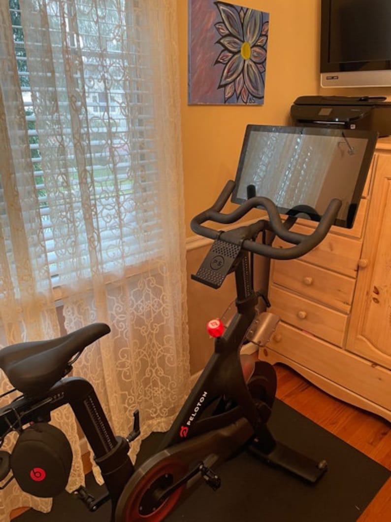 May include: A black stationary exercise bike with a touchscreen display and a red "Peloton" logo. The bike is in a home setting with a window and a dresser in the background.