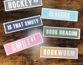 May include: A collection of rectangular signs with white borders and text. The signs feature phrases like "HOCKEY RD," "CLASSICS," "IS THAT SMUT?," "SMUT ST," "BOOK DRAGON," and "BOOKWORM." The signs are in various colors, including blue, pink, green, and tan.