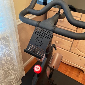 May include: Black bike with a phone holder attached to the handlebars. The phone holder has a black and white grid pattern and the letters "PS" in a circle. The bike has a red knob with a white "P" on it.