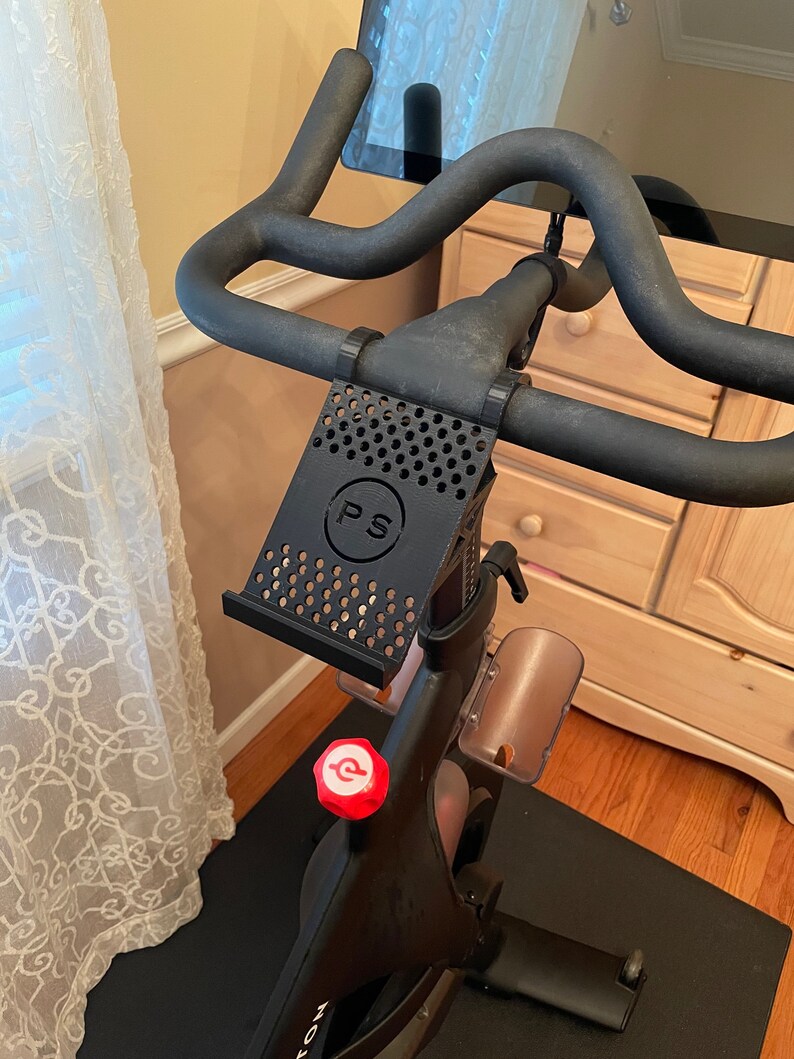 May include: Black bike with a phone holder attached to the handlebars. The phone holder has a black and white grid pattern and the letters "PS" in a circle. The bike has a red knob with a white "P" on it.