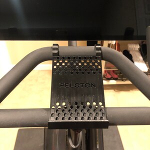 May include: Black 3D printed phone holder with a perforated design and the word "PELOTON" printed on it. The holder is attached to a black exercise bike handlebar.