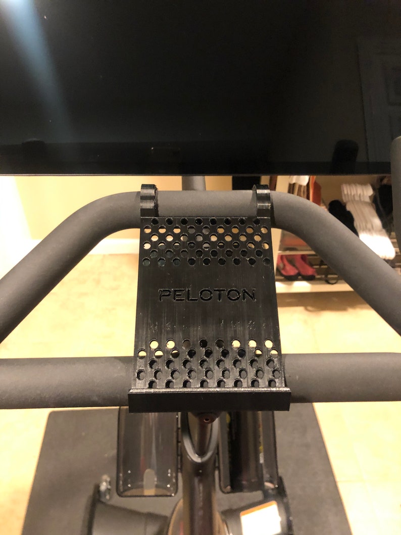 May include: Black 3D printed phone holder with a perforated design and the word "PELOTON" printed on it. The holder is attached to a black exercise bike handlebar.
