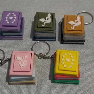 Może przedstawiać: Breloki w kształcie miniaturowych książek w różnych kolorach, w tym fioletowym, zielonym i żółtym. Każda książka ma inny wzór na okładce, np. serce lub kwiatowy wzór. Breloki są przymocowane do srebrnych kółek.