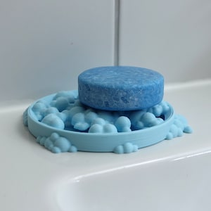 Op de afbeelding: Een lichtblauwe zeepbak in de vorm van wolken met een ronde, lichtblauwe zeep er bovenop.