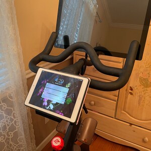 May include: A black stationary exercise bike with a tablet mounted on the handlebars. The tablet screen shows the time 4:29. A larger screen is mounted above the handlebars.