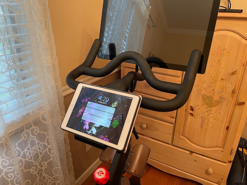 May include: A black stationary exercise bike with a tablet mounted on the handlebars. The tablet screen shows the time 4:29. A larger screen is mounted above the handlebars.