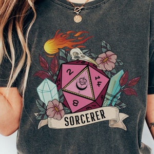 Puede incluir: Una camiseta gris oscuro con un gr&aacute;fico de dado de 20 caras rosa y blanco, rodeado de flores, cristales y llamas. El texto "SORCERER" est&aacute; impreso debajo del dado.