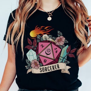 Puede incluir: Camiseta negra con un gr&aacute;fico de dados rosa y blanco, detalles florales y el texto "SORCERER".