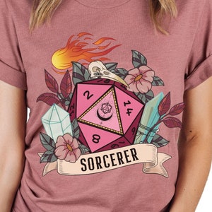 Puede incluir: Una camiseta rosa, roja y blanca con un gr&aacute;fico de un dado rosa de veinte caras con llamas, flores y una calavera. El texto "SORCERER" est&aacute; impreso en una pancarta debajo del dado.