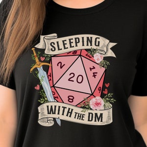 May include: Black t-shirt with a pink and white 20-sided die with a sword and flowers. The text "Sleeping with the DM" is printed on the shirt.