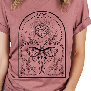 May include: A dusty rose t-shirt with a black graphic design featuring a moth, a twenty-sided die, and a crescent moon.
