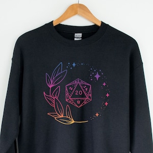 Op de afbeelding: Zwart sweatshirt met ronde hals met een kleurrijke illustratie van een D20-dobbelsteen omringd door bladeren en sterren. Het ontwerp loopt over van roze naar oranje en blauw. Het sweatshirt hangt aan een houten hanger.