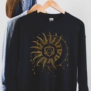 Puede incluir: Sudadera negra con un dise&ntilde;o dorado de sol y luna. El sol presenta una forma geom&eacute;trica en el centro. El dise&ntilde;o incluye puntos y l&iacute;neas doradas. La sudadera est&aacute; colgada en una percha de madera.