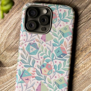 May include: A white phone case with a floral pattern and colorful dice. The dice have different numbers on them, including 1, 4, 7, 16, and 20. The case has a light pink, blue, and green color scheme.