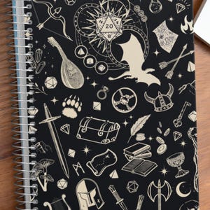 May include: Black spiral-bound notebook with a cream-colored fantasy-themed design. The cover features illustrations of a dragon, dice, sword, and other role-playing game elements. The notebook is on a wooden surface, with dice and a laptop nearby. The text "Dungeon Atelier" is visible.
