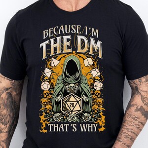 May include: Black t-shirt with a graphic of a hooded figure holding a 20-sided die. The text "Because I'm The DM That's Why" is printed on the shirt.