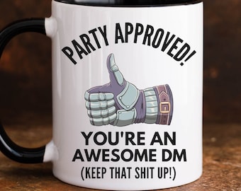 May include: White ceramic coffee mug with a black rim and handle. The mug has a black and white illustration of a thumbs up hand wearing a gauntlet. The text on the mug reads "Party Approved! You're an Awesome DM (Keep that shit up!)"
