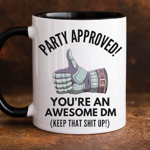 May include: White ceramic coffee mug with a black rim and handle. The mug has a black and white illustration of a thumbs up hand wearing a gauntlet. The text on the mug reads "Party Approved! You're an Awesome DM (Keep that shit up!)"