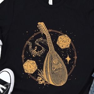 May include: Black t-shirt featuring a gold illustration of a lute, musical notes, and two dice. The dice are numbered 20, 1, 8, and 6. The text on the dice reads "D20".
