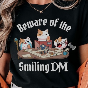 Puede incluir: Camiseta negra con el texto "Beware of the Smiling DM" y una ilustración de dibujos animados de gatos jugando a un juego de mesa. Los gatos tienen diferentes expresiones alrededor de una mesa con dados, mapas y miniaturas. El logo de Dungeon Atelier está en la parte inferior derecha.