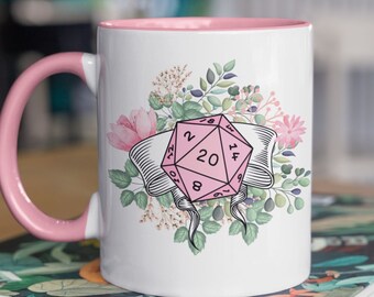 May include: White ceramic mug with a pink rim and handle. The mug features a pink 20-sided die with floral accents. The die has numbers 1, 2, 4, 6, 8, 10, 12, 14, 16, 18, 20, 19, 17, 15, 13, 11, 9, 7, 5, and 3.