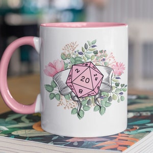 May include: White ceramic mug with a pink rim and handle. The mug features a pink 20-sided die with floral accents. The die has numbers 1, 2, 4, 6, 8, 10, 12, 14, 16, 18, 20, 19, 17, 15, 13, 11, 9, 7, 5, and 3.