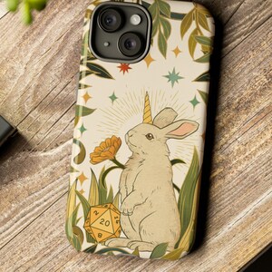 May include: A phone case featuring a whimsical illustration of a rabbit with a unicorn horn, surrounded by flowers, stars, and a D20 die. The case has a cream background with green and gold accents, and the text "Dungeon Atelier" is visible.