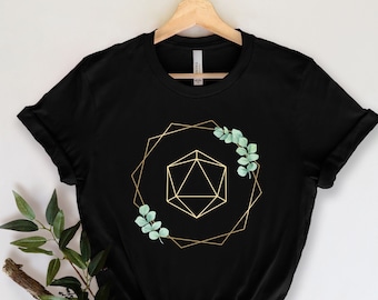 Puede incluir: Camiseta negra con un diseño geométrico dorado, un dado de 20 caras y hojas de eucalipto. El diseño está dentro de un marco geométrico dorado. La camiseta se muestra en una percha de madera.