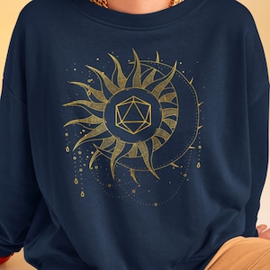 Puede incluir: Sudadera azul marino con un diseño dorado de sol y luna con una forma geométrica en el centro. El sol tiene líneas radiantes y la luna un borde espinoso. El diseño incluye pequeños puntos y gotas.