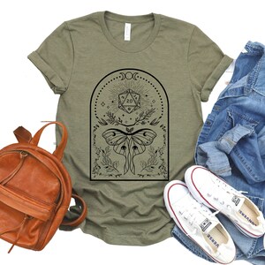 May include: A green t-shirt with a black and white graphic design featuring a moth, a 20-sided die, and a crescent moon. The design is surrounded by a floral border.