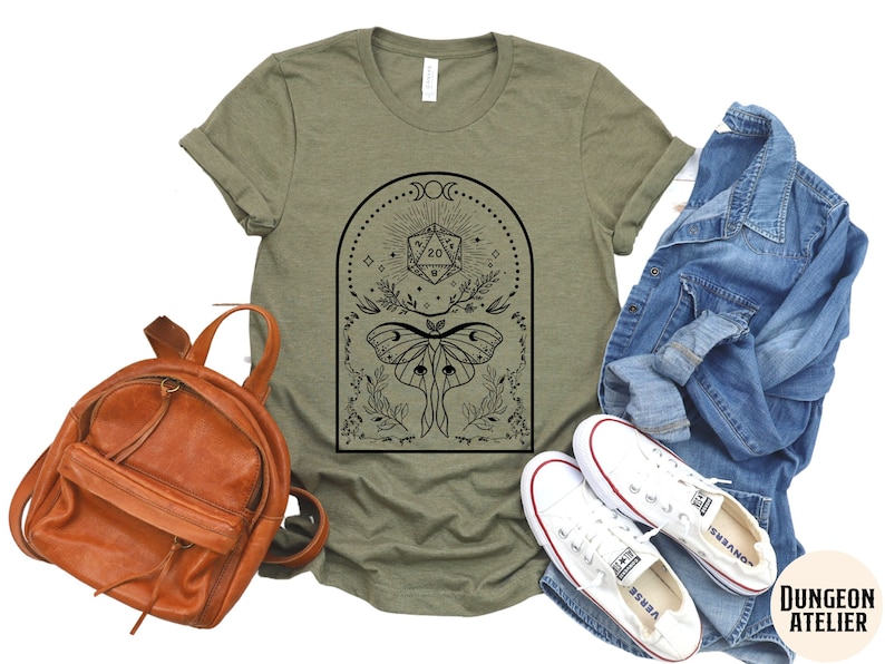 May include: A green t-shirt with a black and white graphic design featuring a moth, a 20-sided die, and a crescent moon. The design is surrounded by a floral border.