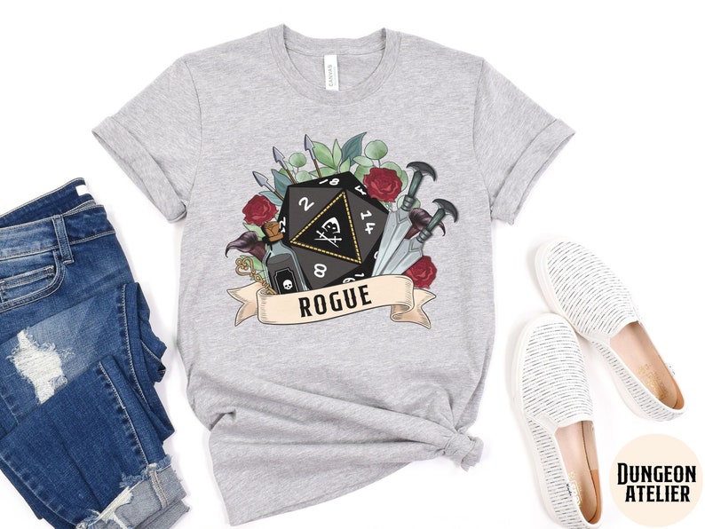 May include: A gray t-shirt with a black and white illustration of a Dungeons and Dragons dice with a skull and crossbones, surrounded by flowers and swords. The text "Rogue" is printed below the dice.