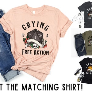 May include: A pink t-shirt with a black and white graphic of a dice with the text "CRYING IS A FREE ACTION".