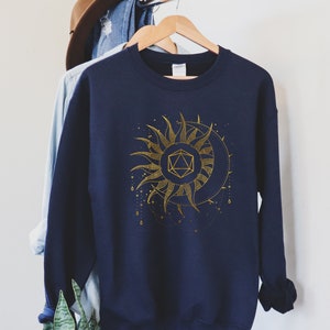 Puede incluir: Sudadera azul marino con un dise&ntilde;o dorado de sol y luna con una forma geom&eacute;trica en el centro. El dise&ntilde;o incluye detalles intrincados y un tema celestial. La sudadera est&aacute; colgada en una percha de madera.