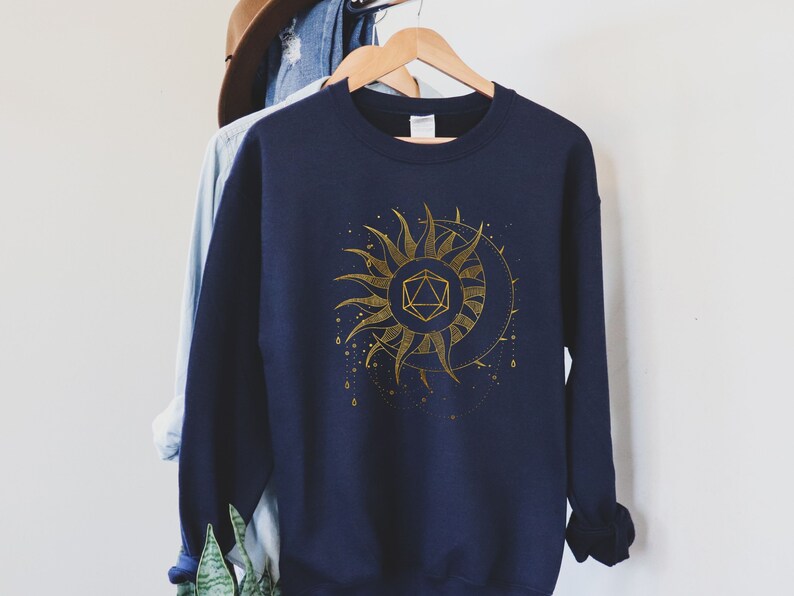 Puede incluir: Sudadera azul marino con un dise&ntilde;o dorado de sol y luna con una forma geom&eacute;trica en el centro. El dise&ntilde;o incluye detalles intrincados y un tema celestial. La sudadera est&aacute; colgada en una percha de madera.