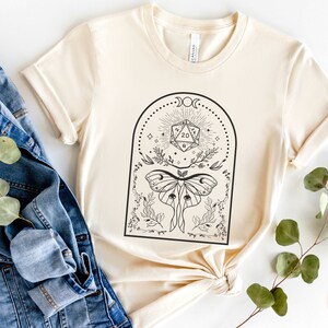 May include: A cream-colored t-shirt with a black and white graphic design featuring a moth, a twenty-sided die, and a crescent moon. The text "Dungeon Atelier" is visible in the bottom right corner of the image.