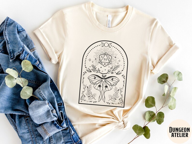 May include: A cream-colored t-shirt with a black and white graphic design featuring a moth, a twenty-sided die, and a crescent moon. The text "Dungeon Atelier" is visible in the bottom right corner of the image.