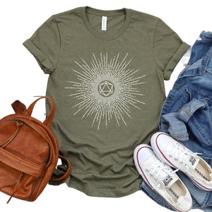 Puede incluir: Una camiseta verde con un gr&aacute;fico blanco de un dado geom&eacute;trico con un dise&ntilde;o de rayos de sol. La camiseta se combina con una mochila de cuero marr&oacute;n, una chaqueta vaquera y zapatillas blancas de lona.