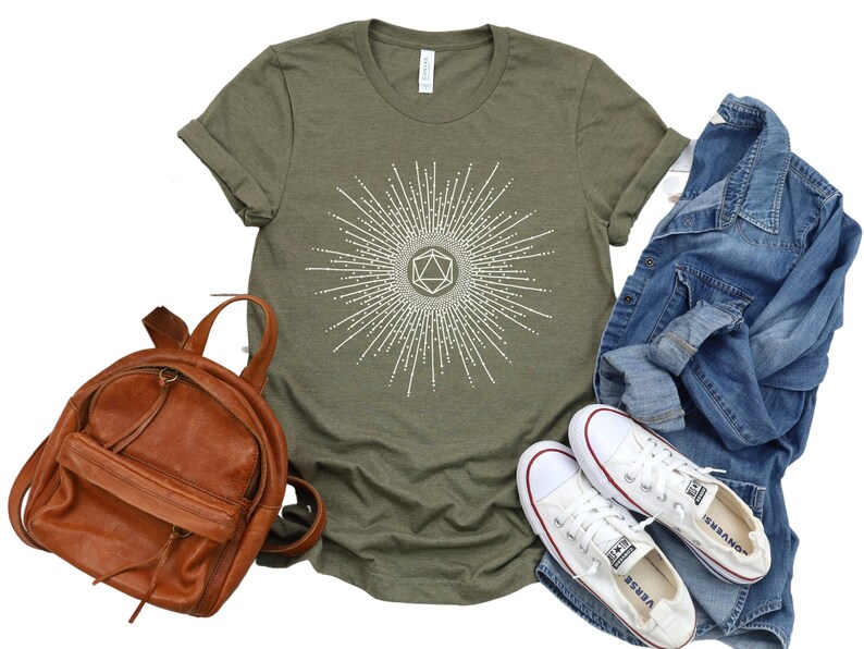 Puede incluir: Una camiseta verde con un gr&aacute;fico blanco de un dado geom&eacute;trico con un dise&ntilde;o de rayos de sol. La camiseta se combina con una mochila de cuero marr&oacute;n, una chaqueta vaquera y zapatillas blancas de lona.