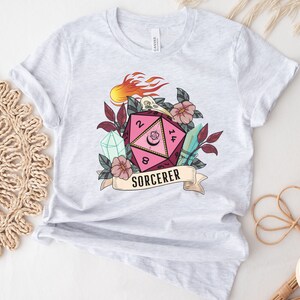 Puede incluir: Una camiseta blanca con un dado rosa y negro de 20 caras con una luna creciente y una calavera. El dado est&aacute; rodeado de flores, cristales y una pancarta que dice "SORCERER".