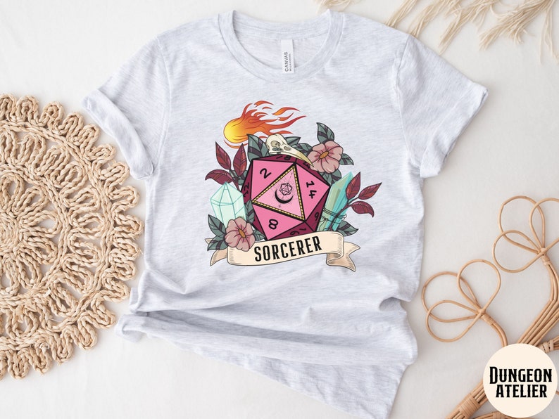 Puede incluir: Una camiseta blanca con un dado rosa y negro de 20 caras con una luna creciente y una calavera. El dado est&aacute; rodeado de flores, cristales y una pancarta que dice "SORCERER".