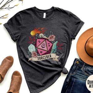 Puede incluir: Una camiseta gris oscuro con un dado rosa y blanco de 20 caras con una luna creciente y estrellas. El dado est&aacute; rodeado de flores, cristales y una calavera con llamas. El texto "SORCERER" est&aacute; en una pancarta debajo del dado.