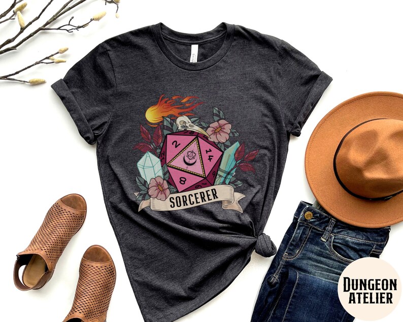 Puede incluir: Una camiseta gris oscuro con un dado rosa y blanco de 20 caras con una luna creciente y estrellas. El dado est&aacute; rodeado de flores, cristales y una calavera con llamas. El texto "SORCERER" est&aacute; en una pancarta debajo del dado.