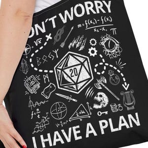 May include: Black tote bag with a white graphic design featuring a D20 die, mathematical equations, and the text "DON'T WORRY I HAVE A PLAN". The bag is held by a person.